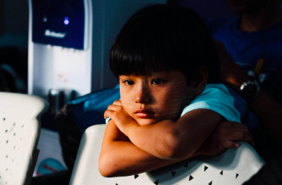 A young child with a solemn expression rests their chin on folded arms on a white chair back.
