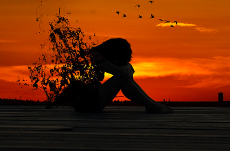 Silhouette of a person sitting alone with head bowed against an orange sunset, birds flying away