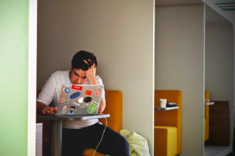Man looking stressed while working on a sticker-covered laptop in a home office setting