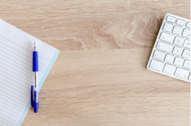 Wooden desk with open notebook, blue pen, and white keyboard viewed from above