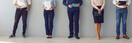Five professionals in business attire standing in a row against a gray wall, holding documents and tablets