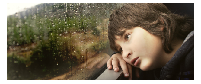 A sad child rests their chin on their hand, gazing out a rain-streaked window at blurred greenery.