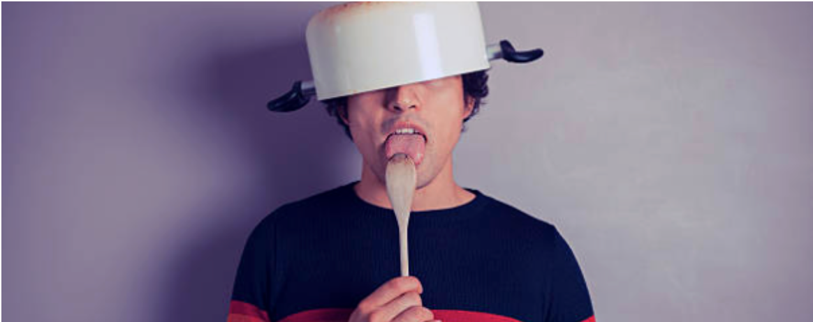 Person wearing a cooking pot upside down on their head and licking a wooden spoon