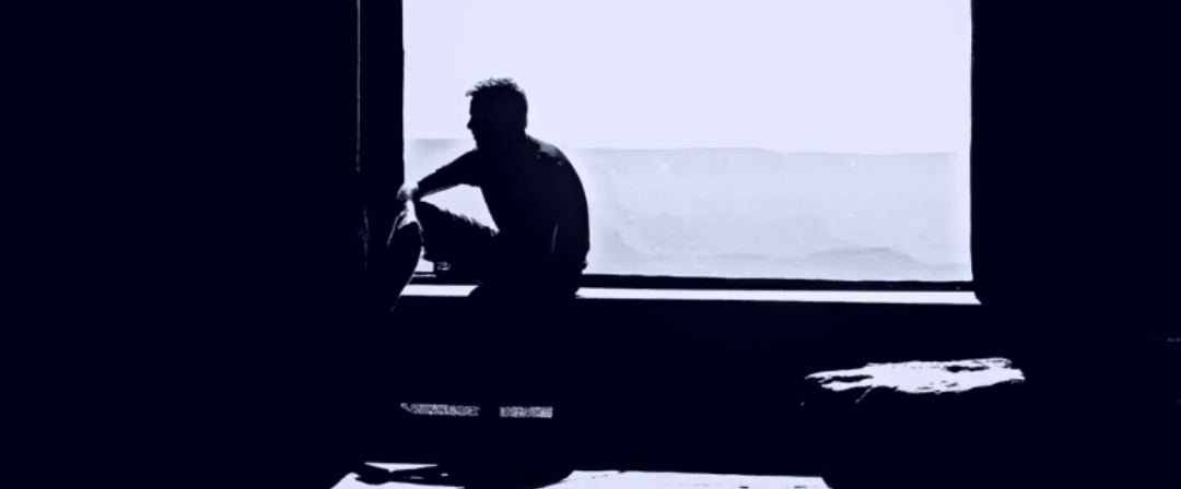 Dark silhouette of a person sitting on a windowsill in an empty room, backlit by pale light