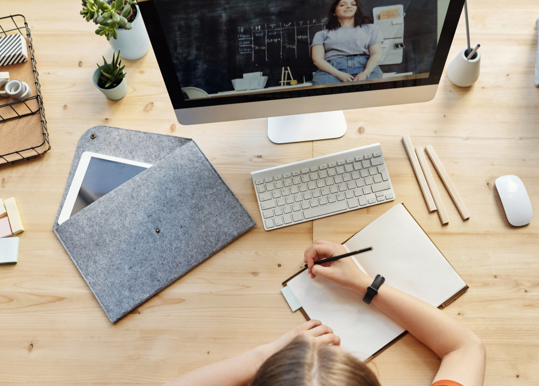 Child writing in notebook at desk with computer showing video lesson, keyboard, tablet, and plants